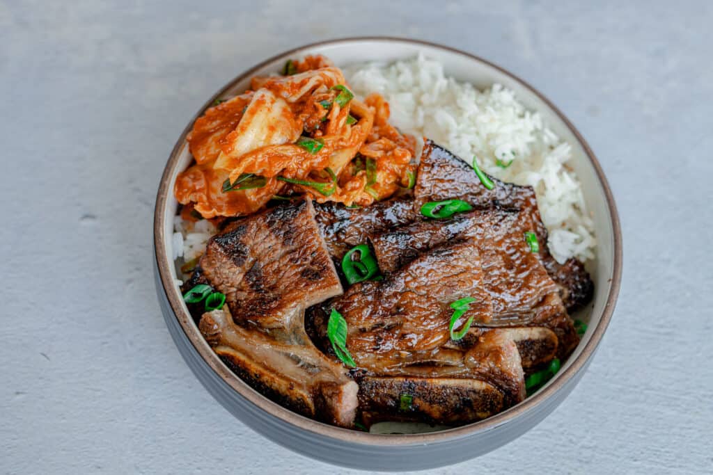 Cut up grilled kalbi served in a bowl with white rice and kim chi
