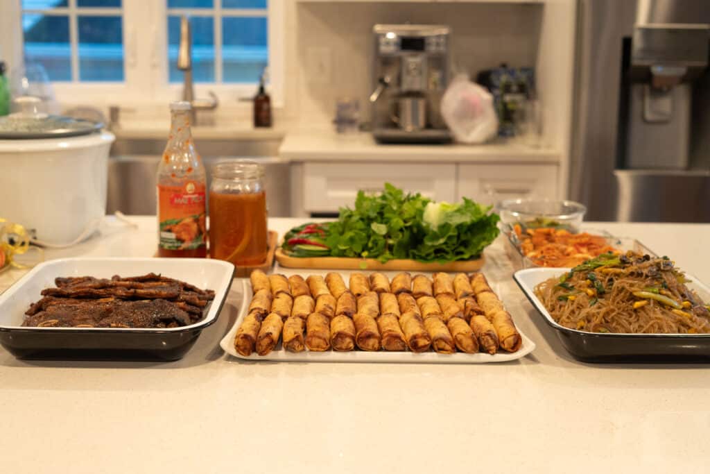 Kalbi served at a party buffet style with egg rolls, noodles, kim chi and other various items.